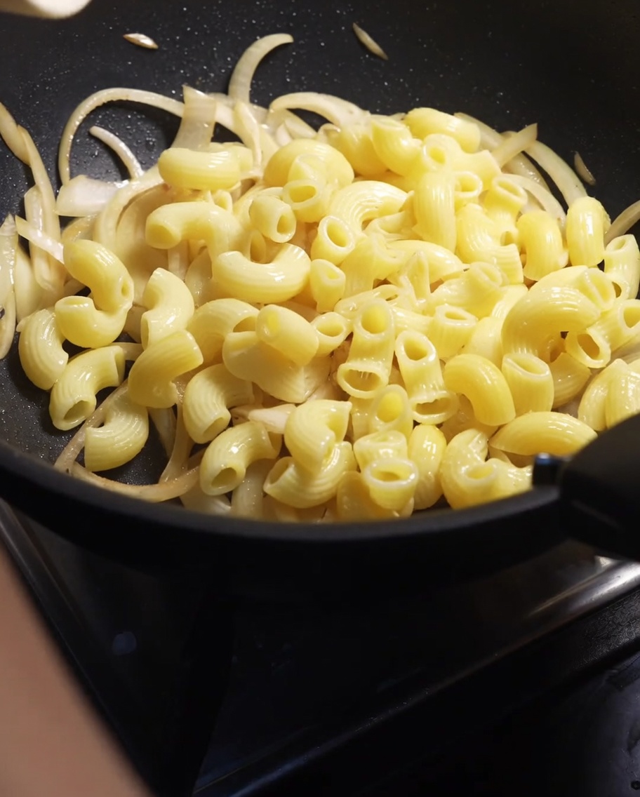 Cooked macaroni elbows being poured into a pan with softened sliced onions.