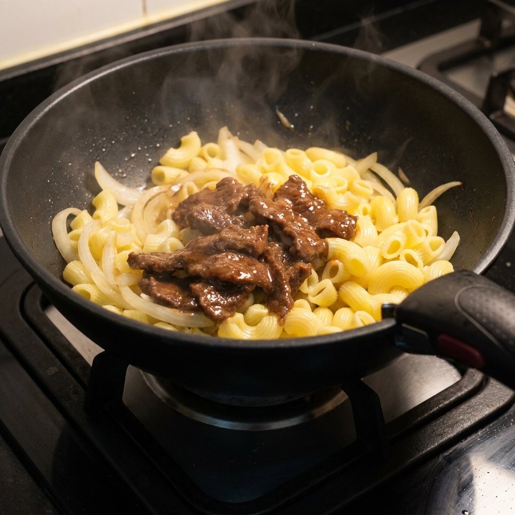 Seared slices of marinated beef being added back into the skillet on top of the macaroni and onions.