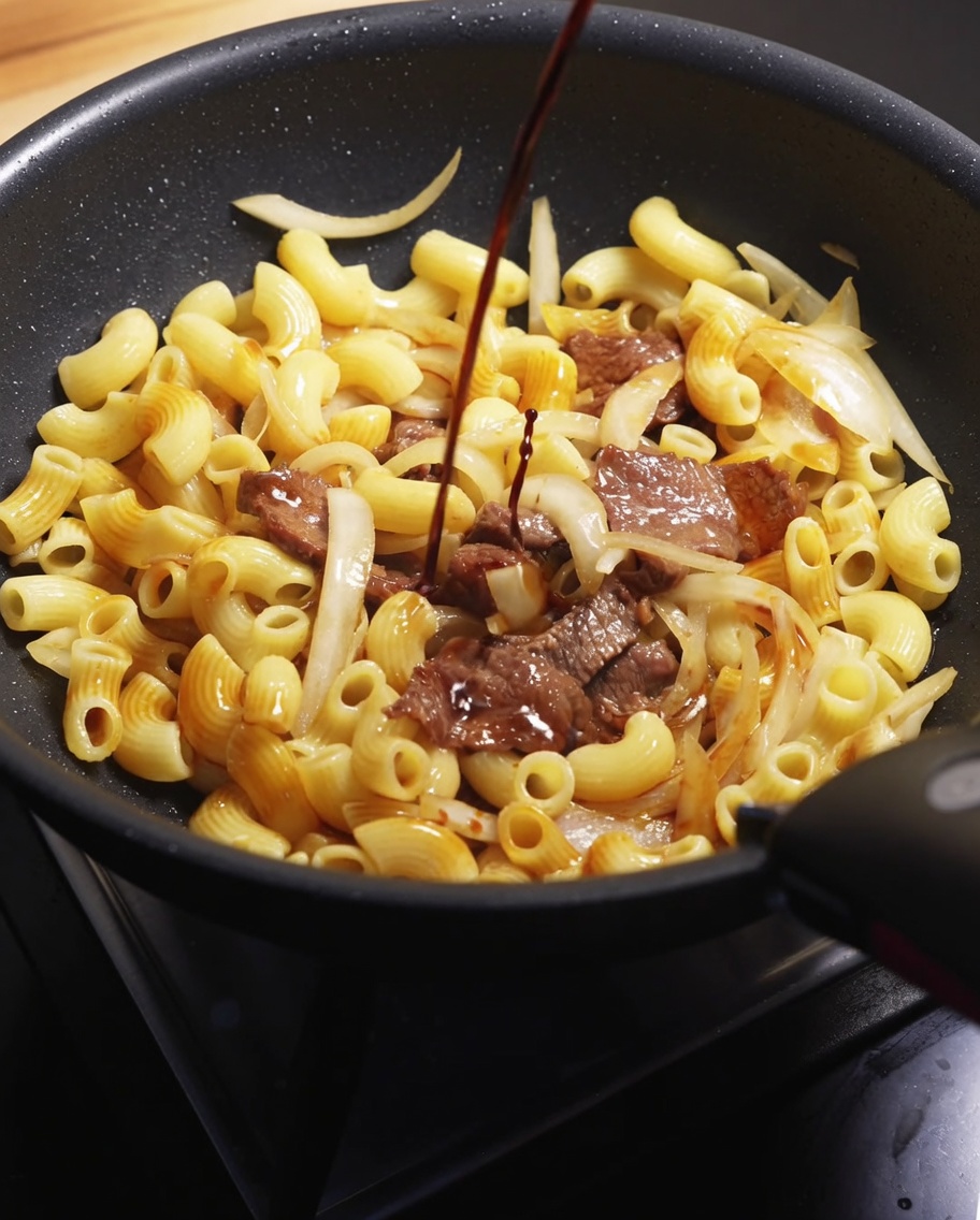 A stream of dark soy sauce being poured over the beef and macaroni mixture in a hot skillet.