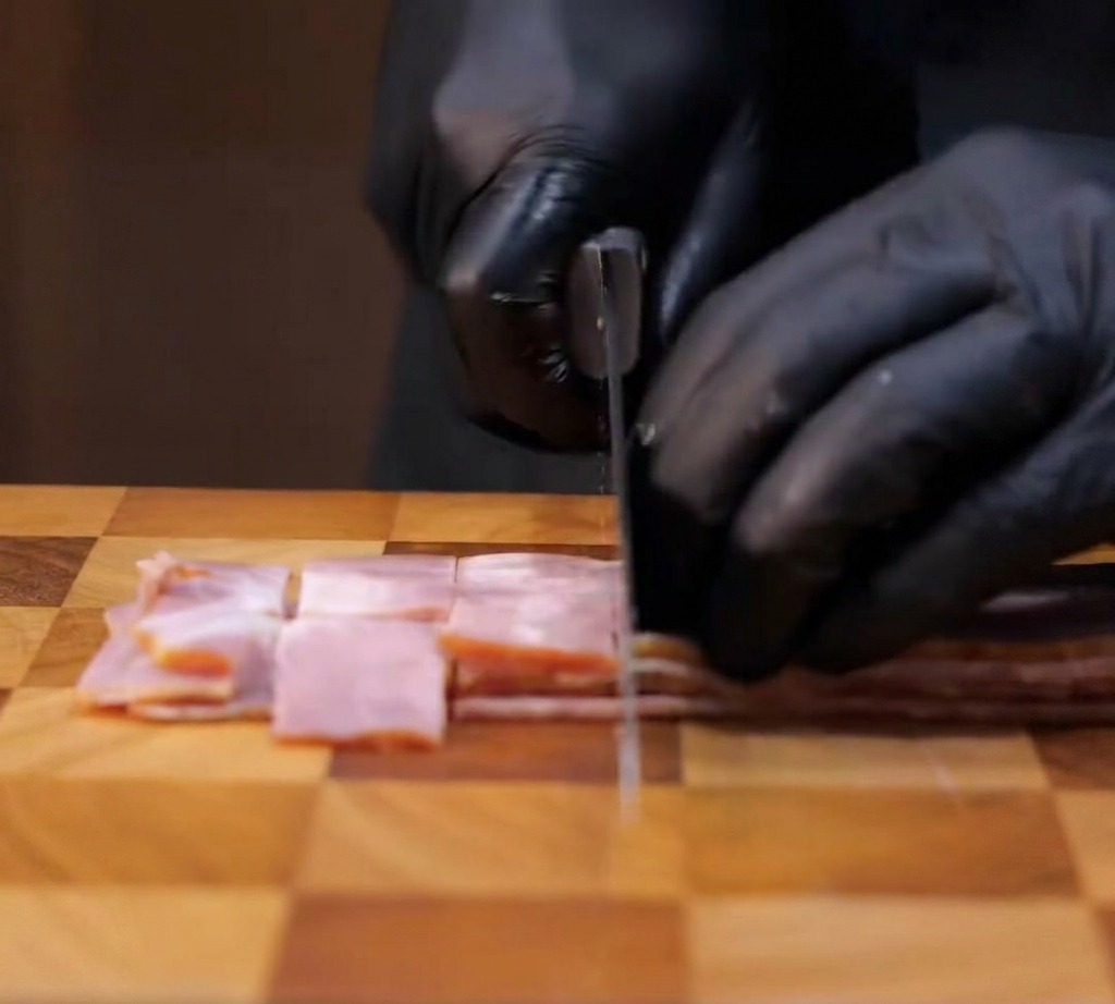 Bacon slices being sliced into square, bite-sized chunks on a wooden cutting board.