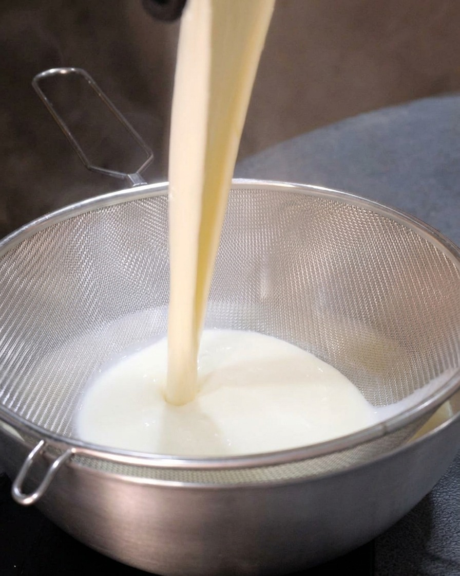 The creamy white soup base being poured through a metal sieve, resulting in a smooth, lump-free liquid in the bowl below.