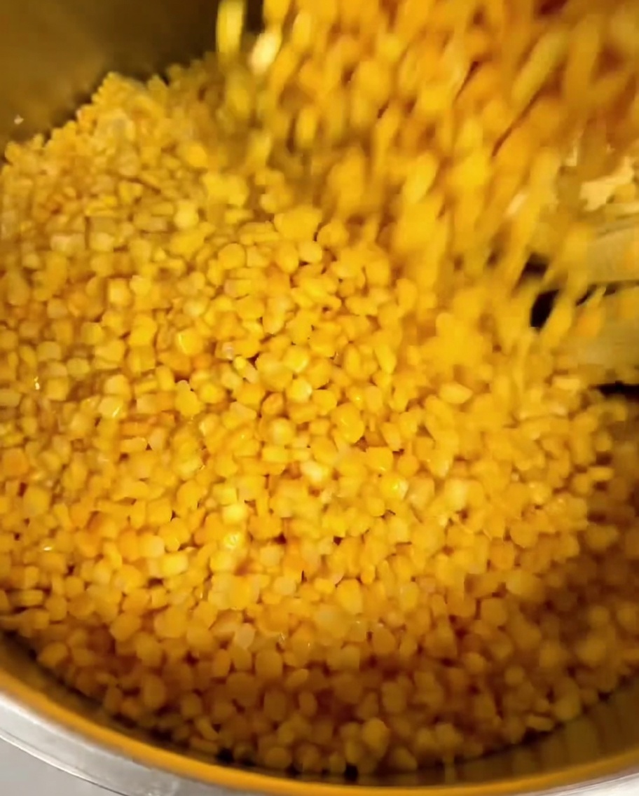 Canned corn kernels being poured over fresh corn kernels in a large metal pot.