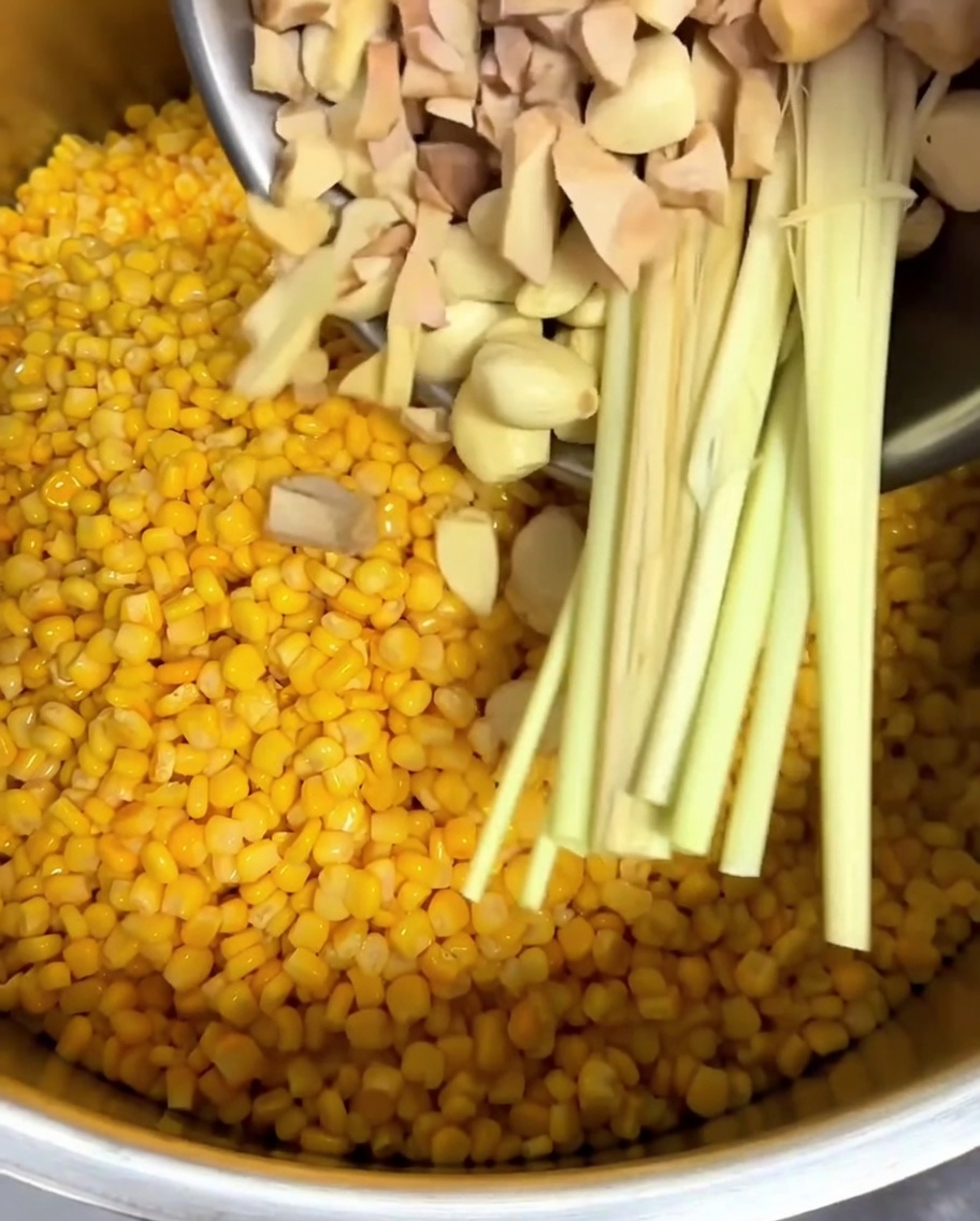 Chopped galangal, whole garlic cloves, and fresh lemongrass stalks being added to a pot of corn kernels.