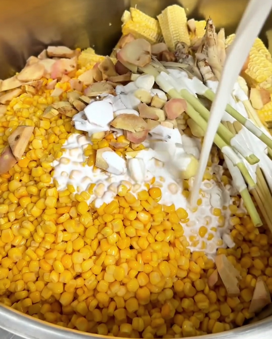 White coconut milk being poured over corn, aromatics, and bare corn cobs in a metal pot.