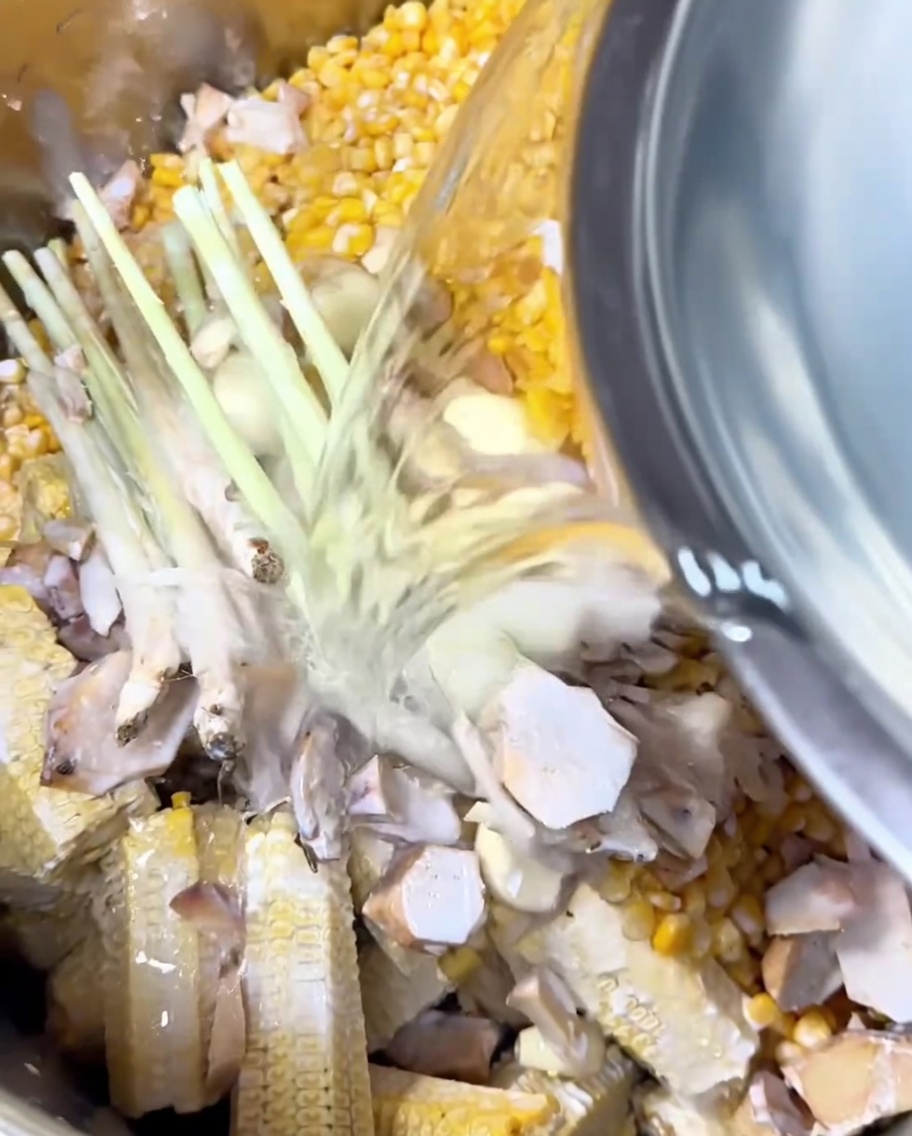 Clear water being poured from a metal bowl into a pot filled with corn, aromatics, and coconut milk.