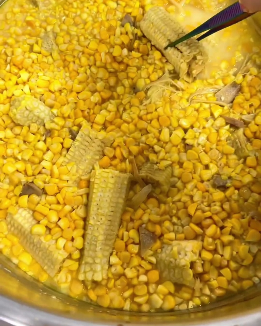 Tongs removing lemongrass and a bare corn cob from a pot of simmering corn soup.