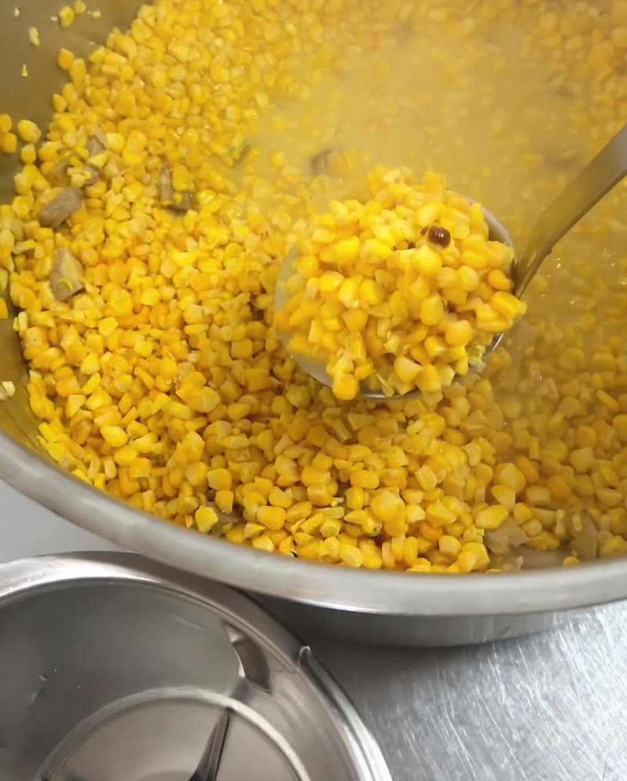 A metal ladle scooping soft, cooked corn kernels from the soup pot.