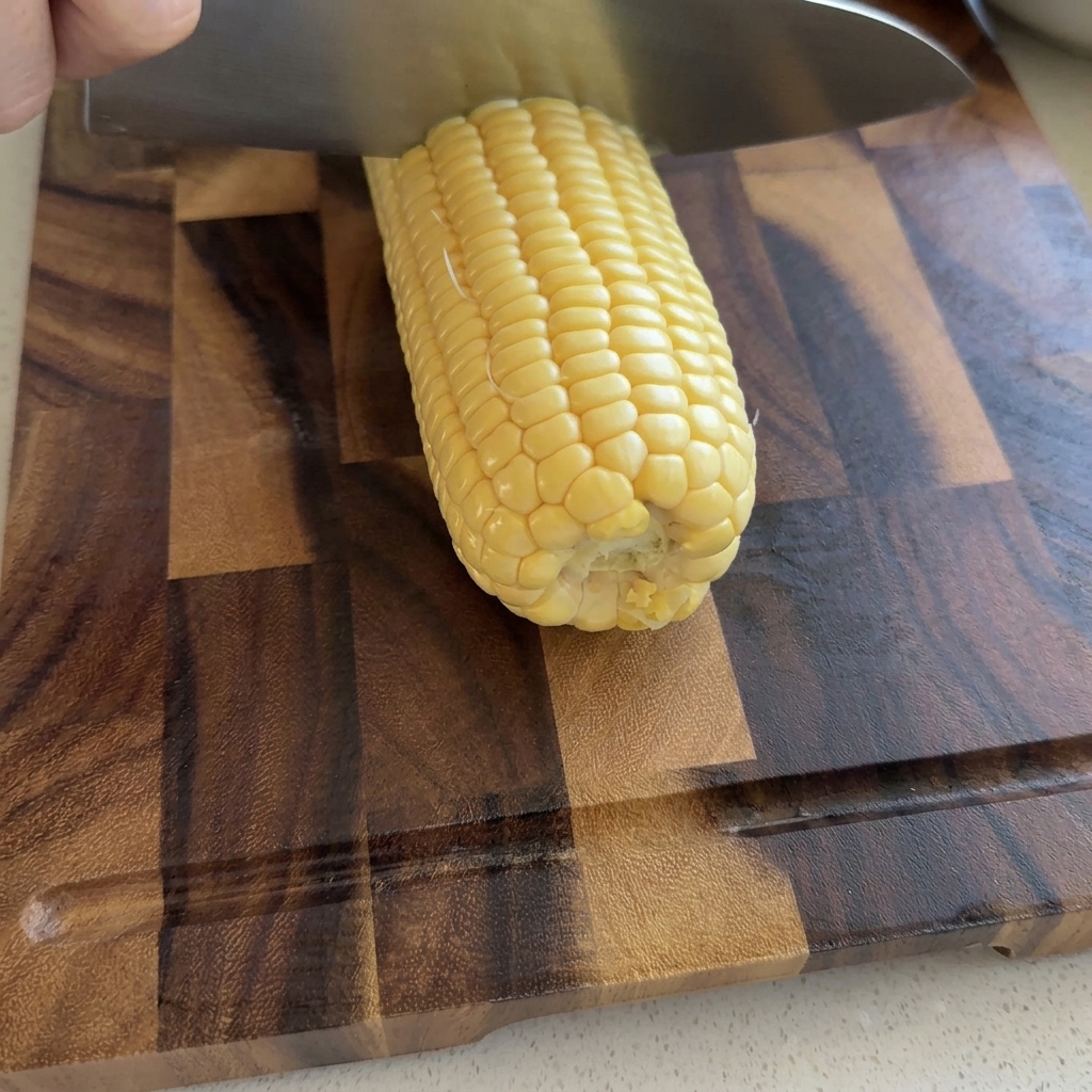A hand using a large chefs knife to slice raw yellow corn kernels off a cob on a wooden cutting board.