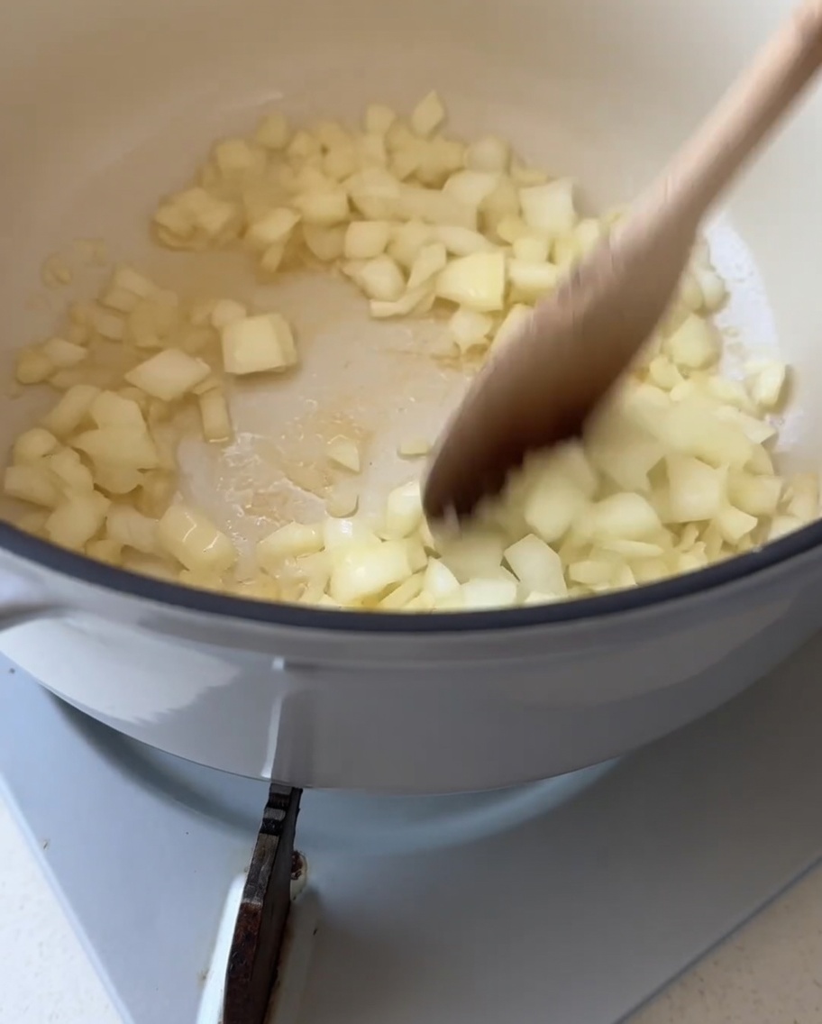 A wooden spoon stirring diced white onions inside a white pot with melted butter.