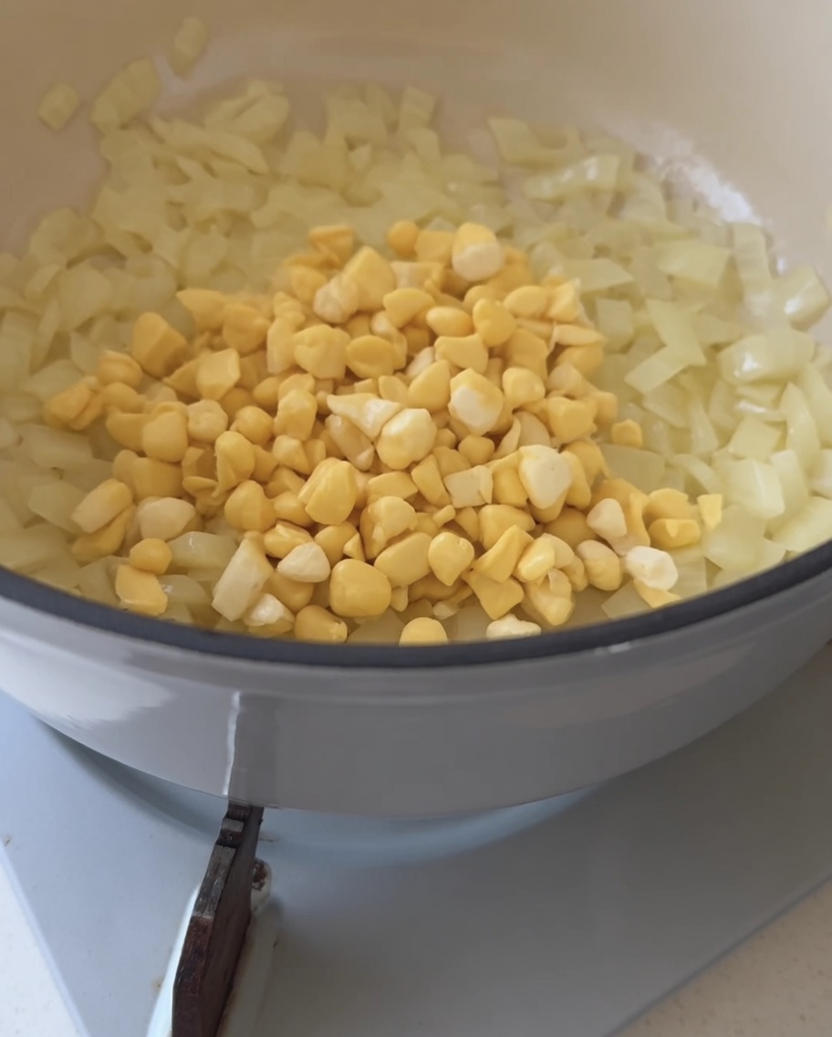 Fresh yellow corn kernels being poured into a pot containing translucent sautéed onions.