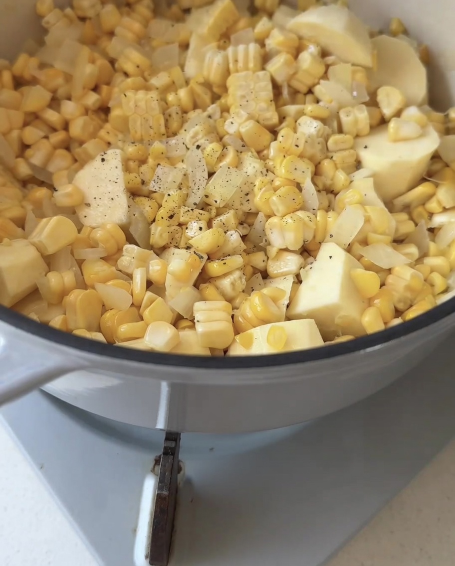 Black pepper being freshly ground over a mixture of corn, onion, and sweet potato in a cooking pot.