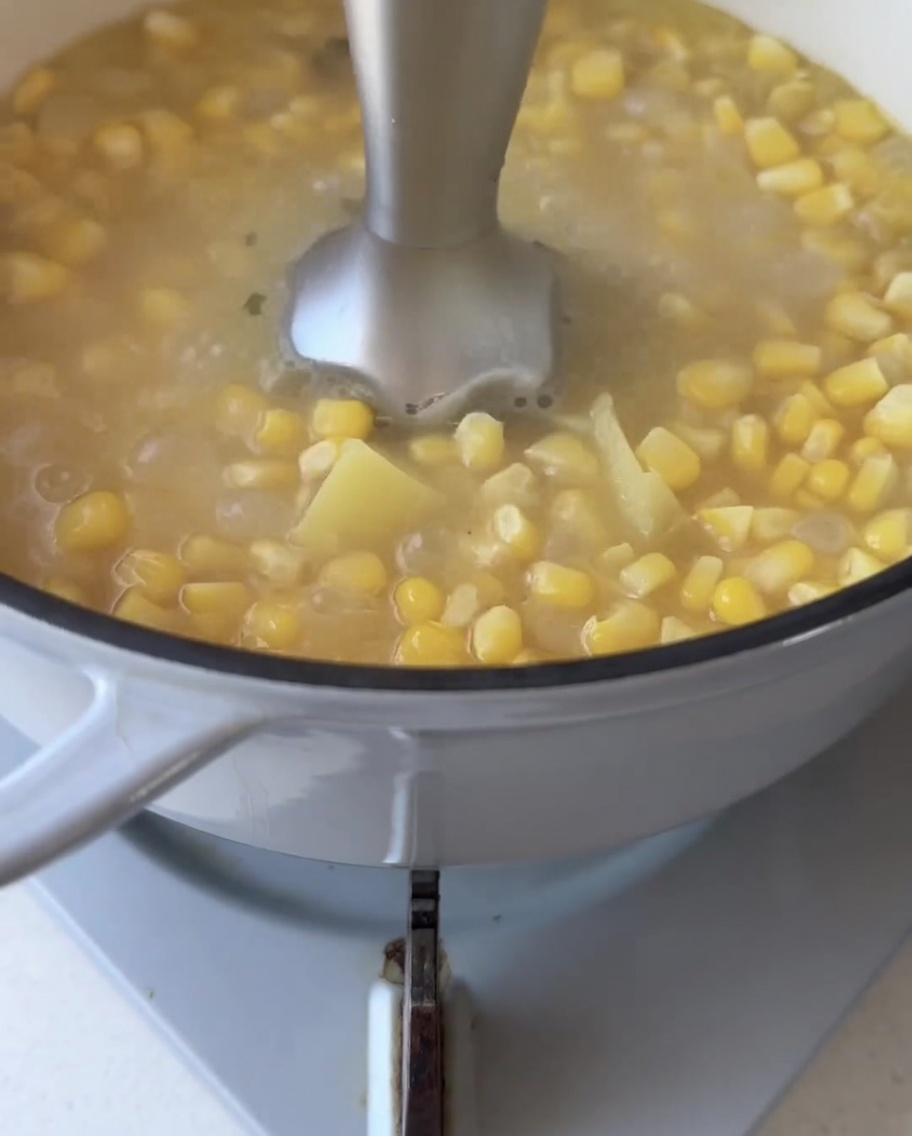 An immersion blender submerged in a pot of simmering corn kernels and sweet potato chunks, blending them into a smooth liquid.