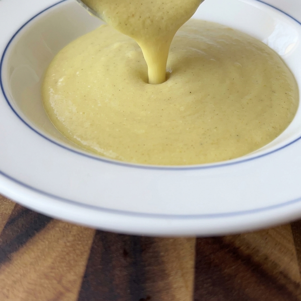 Thick, creamy yellow corn soup being poured from a ladle into a white bowl with a blue rim.