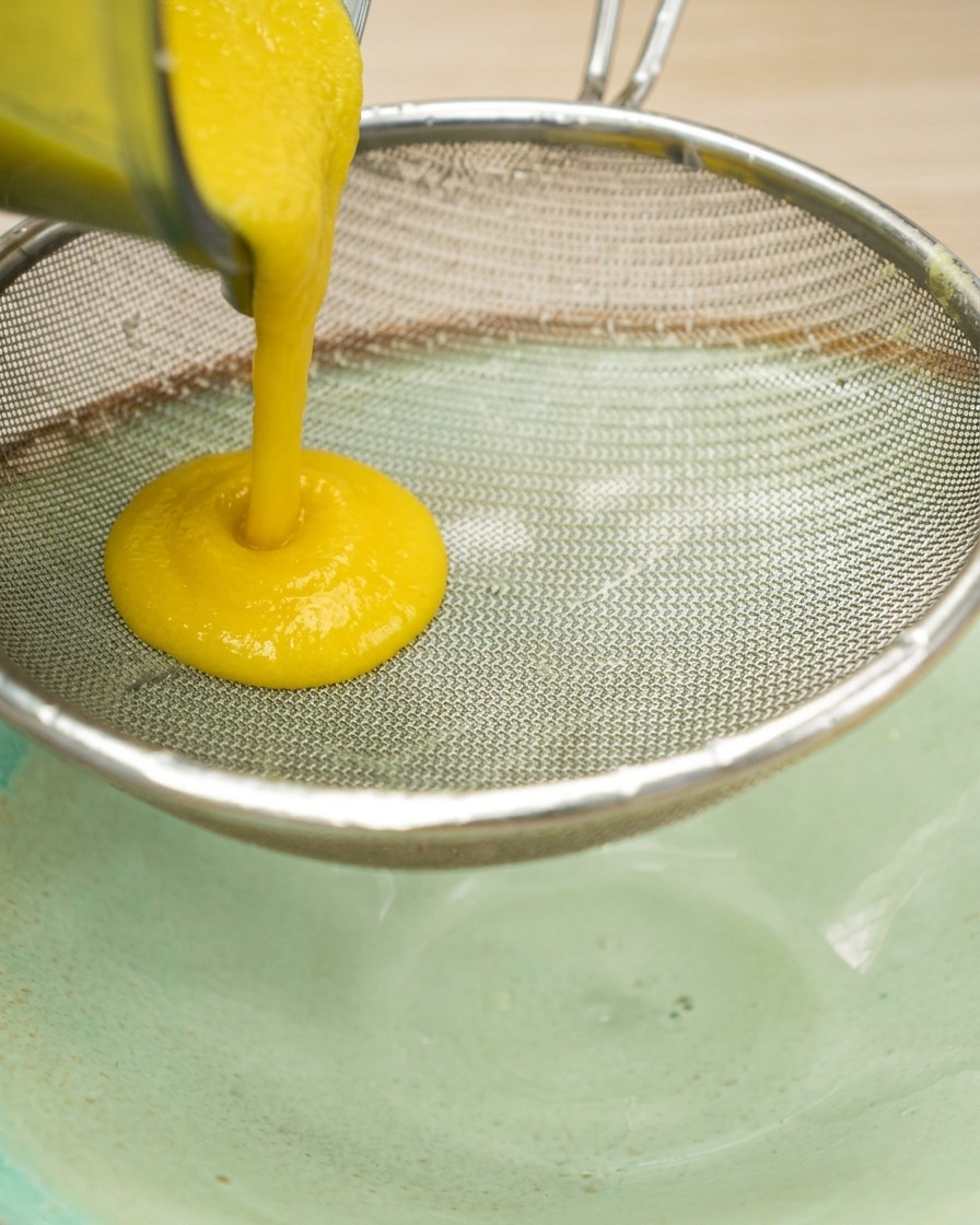 Thick yellow corn puree being strained through a stainless steel fine-mesh sieve into a bowl.