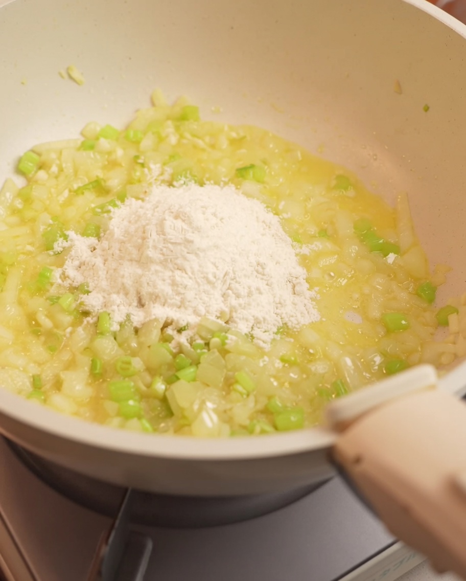 Flour being stirred into the sautéed onion and celery mixture in a pan.