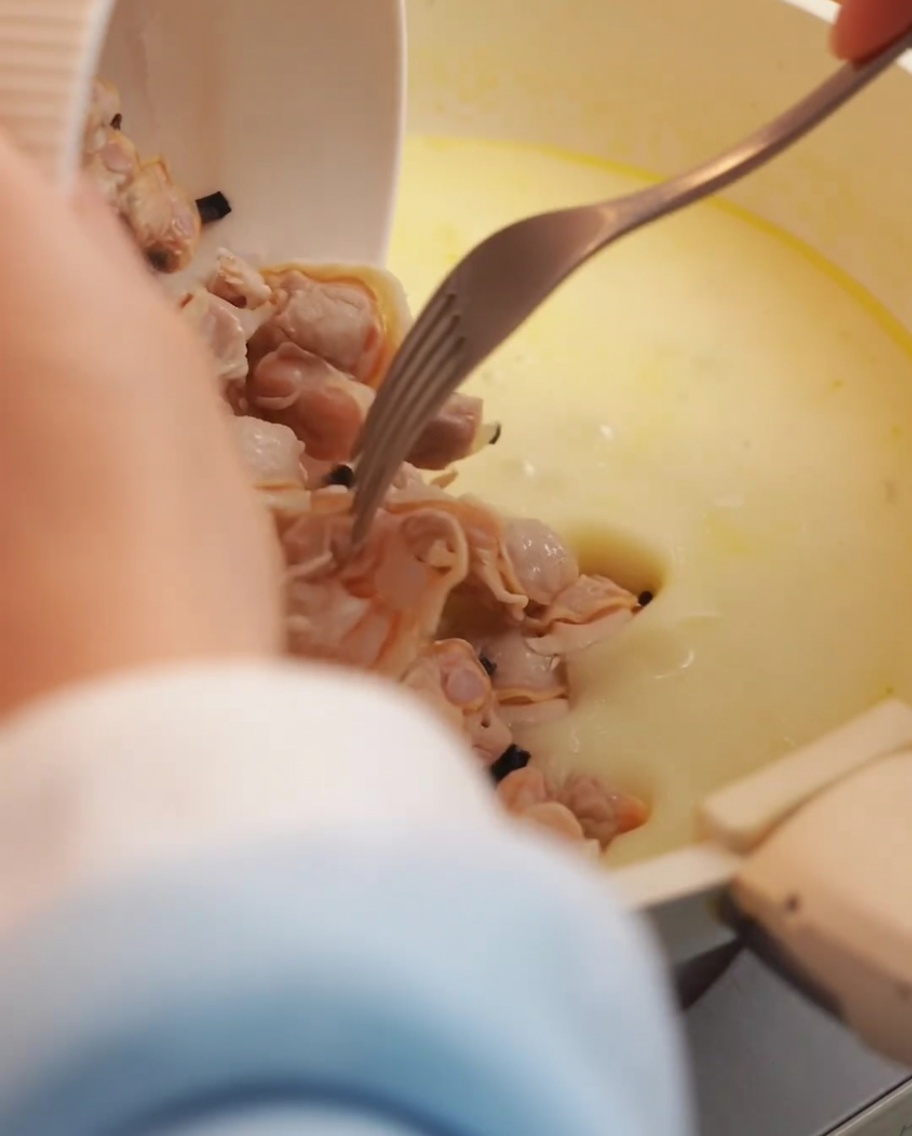 Fresh clam meat and crispy bacon pieces being added to the creamy soup base in the pot.
