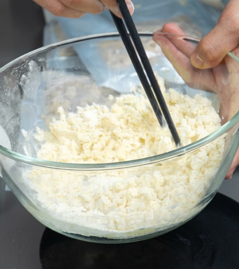 A bowl containing a flour and egg mixture being stirred with chopsticks to form crumbles.