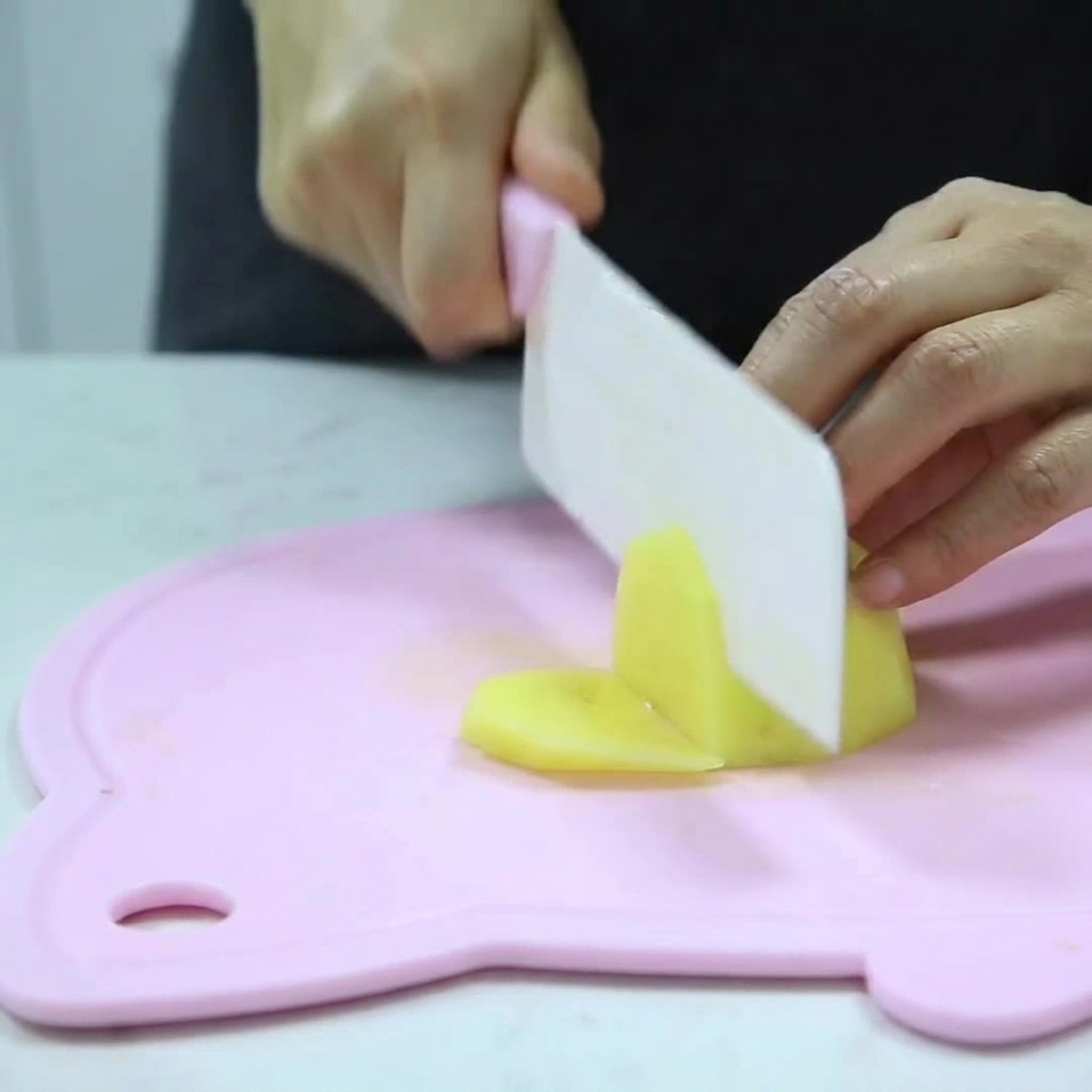 Hands holding a knife, cutting a peeled potato into small cubes on a pink cutting board.