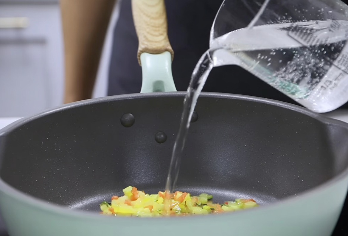 Water being poured from a measuring cup into a pan with sautéed vegetables, preparing to incorporate the thickened milk mixture.