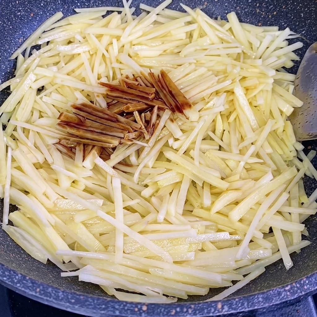Soy sauce and oyster sauce being poured over the stir-frying potato strips in a wok.