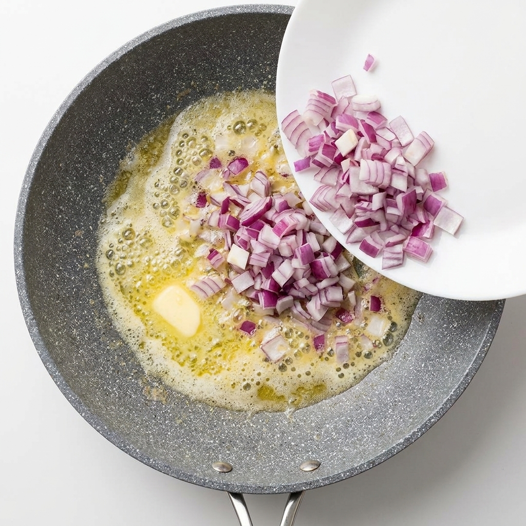 Diced red onions being poured from a white plate into a bubbling pan of melted butter.