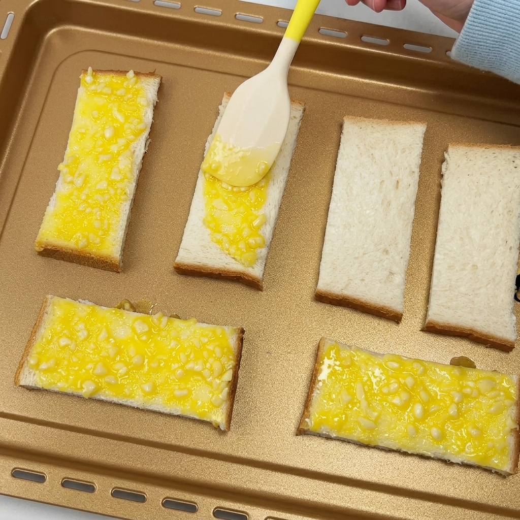 A spatula spreading garlic butter over slices of bread on a baking tray.