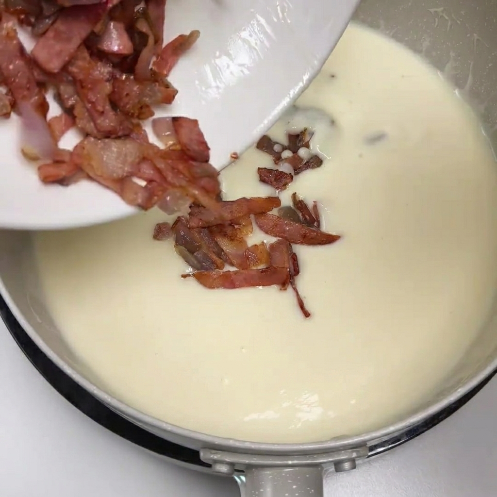 Sautéed bacon and onions being scraped from a plate into the creamy potato soup.