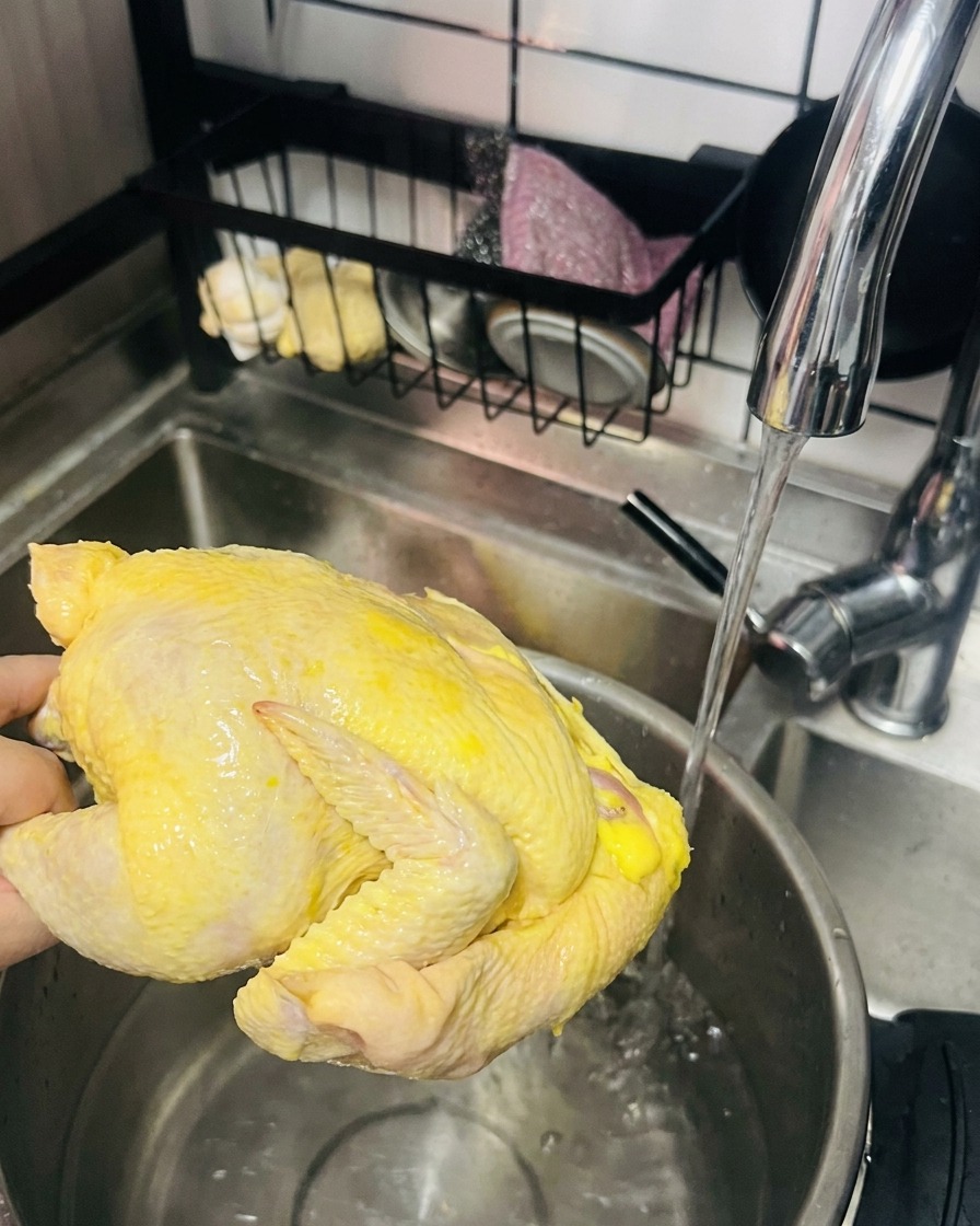 A hand holding a whole raw yellow chicken under a running faucet, ready to be placed in a pot of hot water.