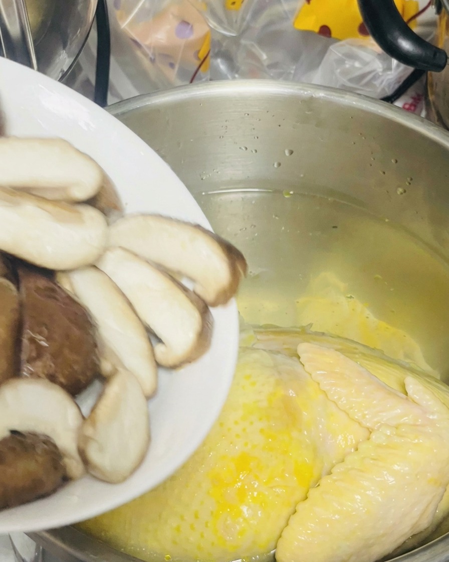 Sliced shiitake mushrooms being poured from a white plate into a pot containing a whole chicken in water.