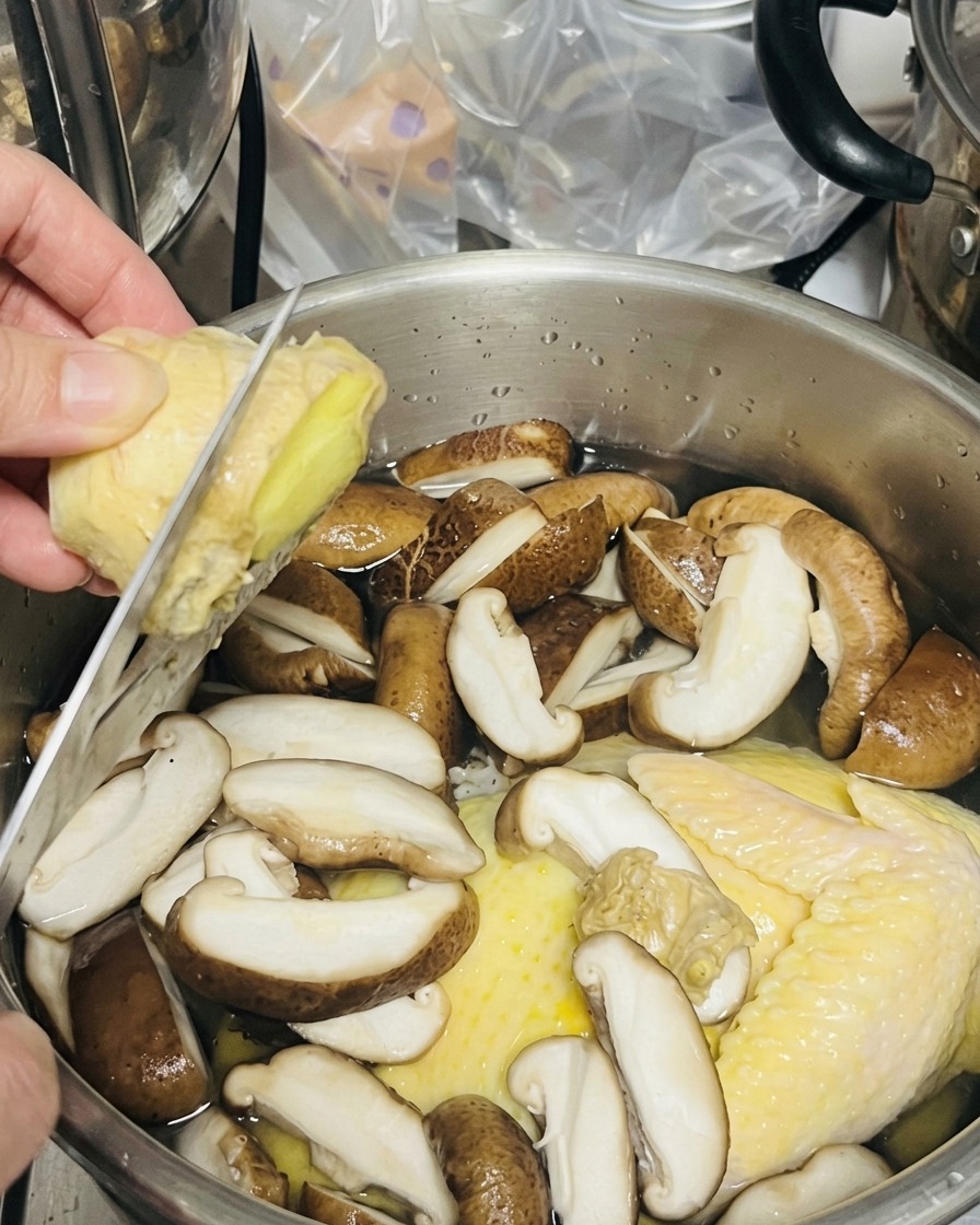 A person using a knife to slice a chunk of fresh ginger directly over a pot of chicken and mushrooms.
