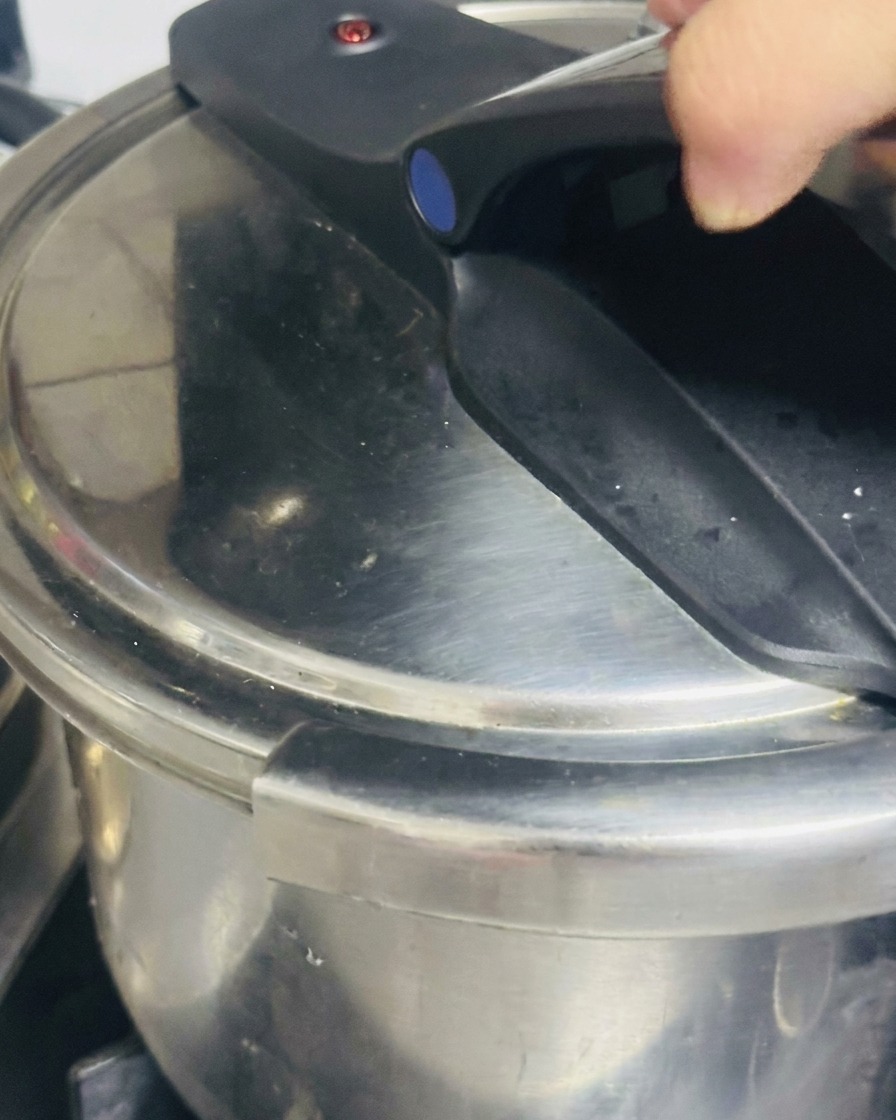 A hand firmly pressing down and locking the black handle of a stainless steel pressure cooker lid.
