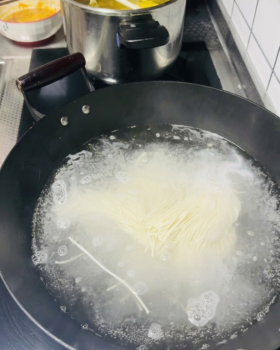 A bundle of thin, raw white noodles being dropped into a black wok filled with vigorously boiling water.
