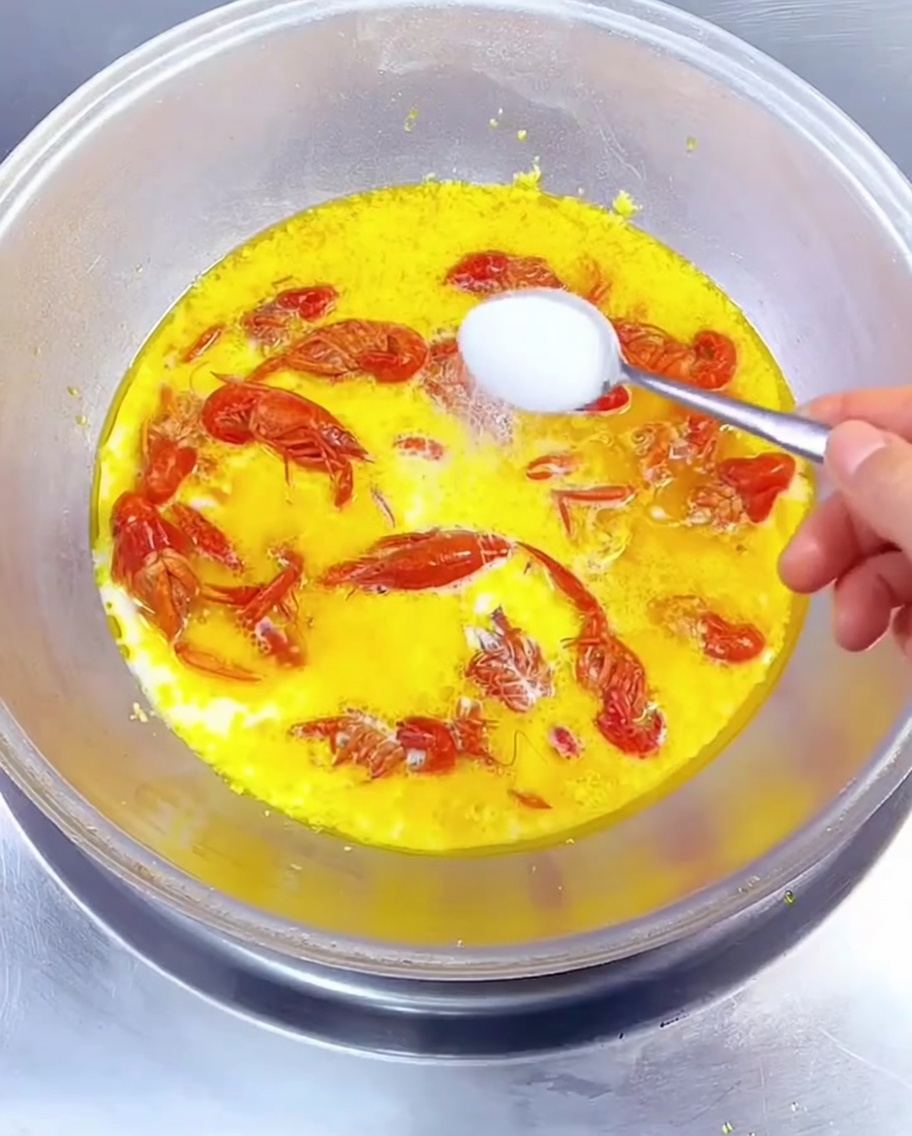 Close-up of a spoon adding white seasoning into a simmering wok of golden milk-based sauce with red crayfish.