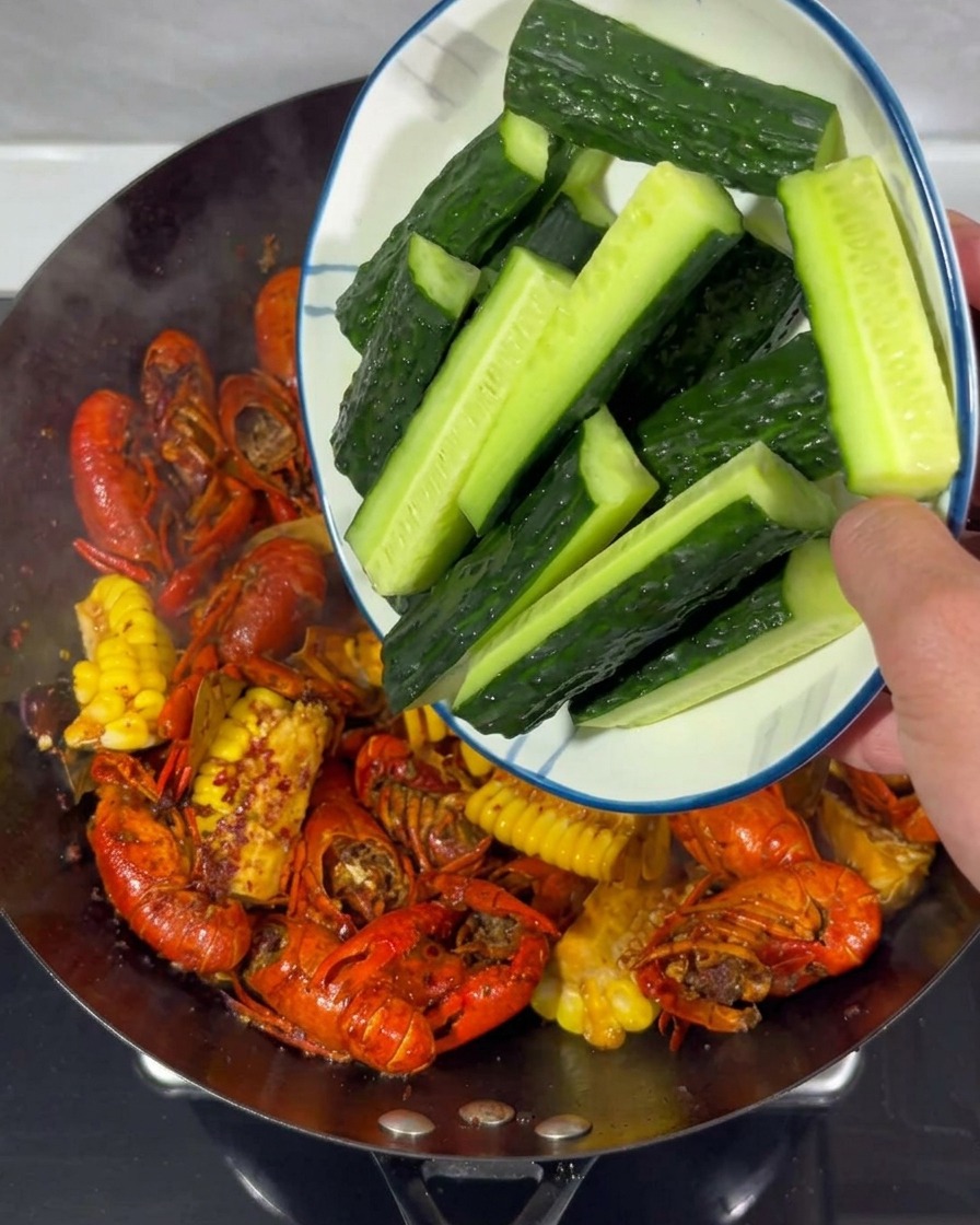 Fresh cucumber sticks being poured from a white plate into a wok containing cooked crayfish and corn.