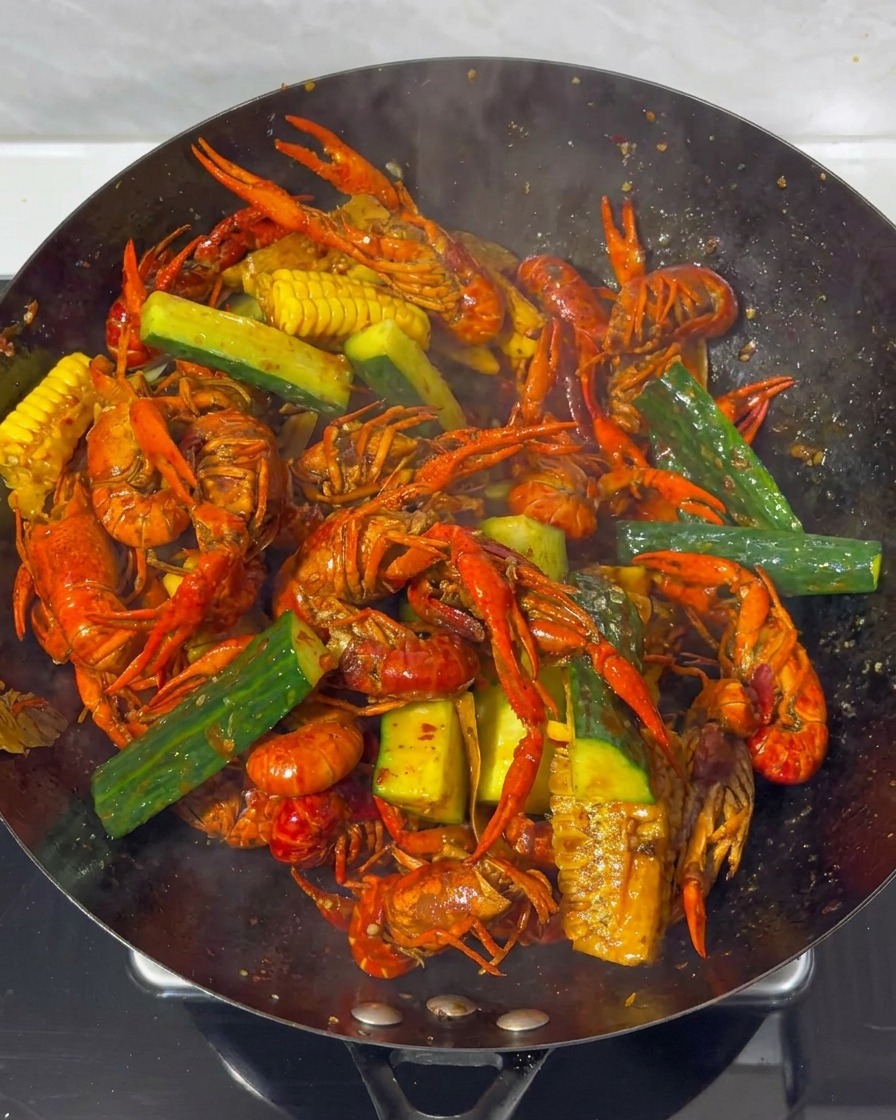 A close-up of crayfish, corn, and cucumber pieces being tossed together in a large wok with a rich, dark red sauce.