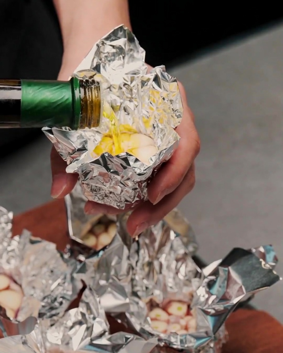 Olive oil being poured from a green bottle onto the exposed cloves of a garlic head resting in aluminum foil.