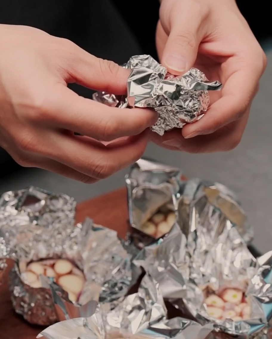 Garlic bulb wrapped in aluminum foil on a wooden cutting board.