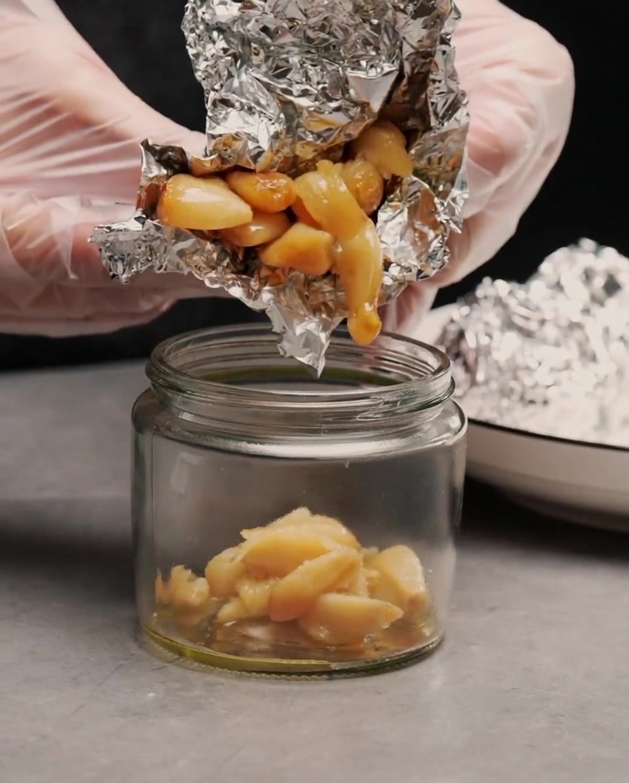 Gloved hands squeezing soft, golden roasted garlic cloves out of aluminum foil and into a small glass jar.