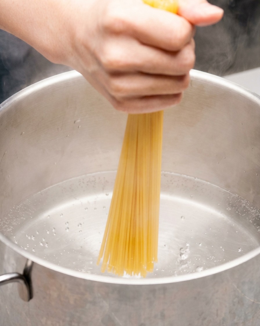 A hand pushing a bundle of dry spaghetti straight down into a stainless steel pot of boiling water.
