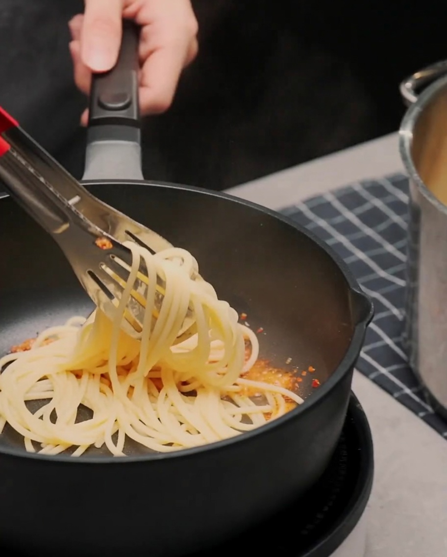 Cooked spaghetti being tossed with tongs in a pan with garlic and chili oil.