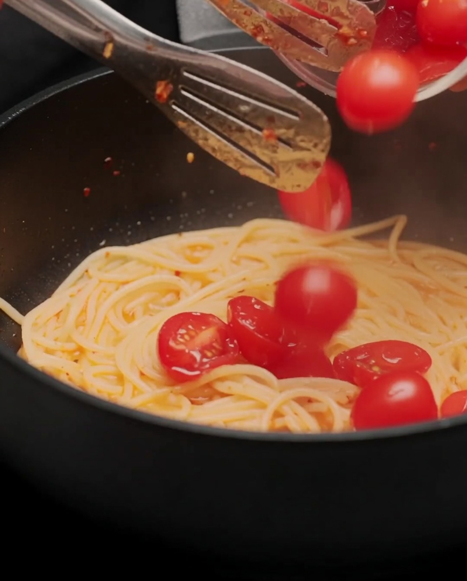 Halved cherry tomatoes being added to the pan of spaghetti and stir-fried.