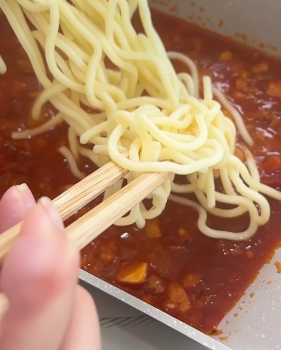 Spaghetti noodles being tossed with tomato meat sauce in a pan using chopsticks.