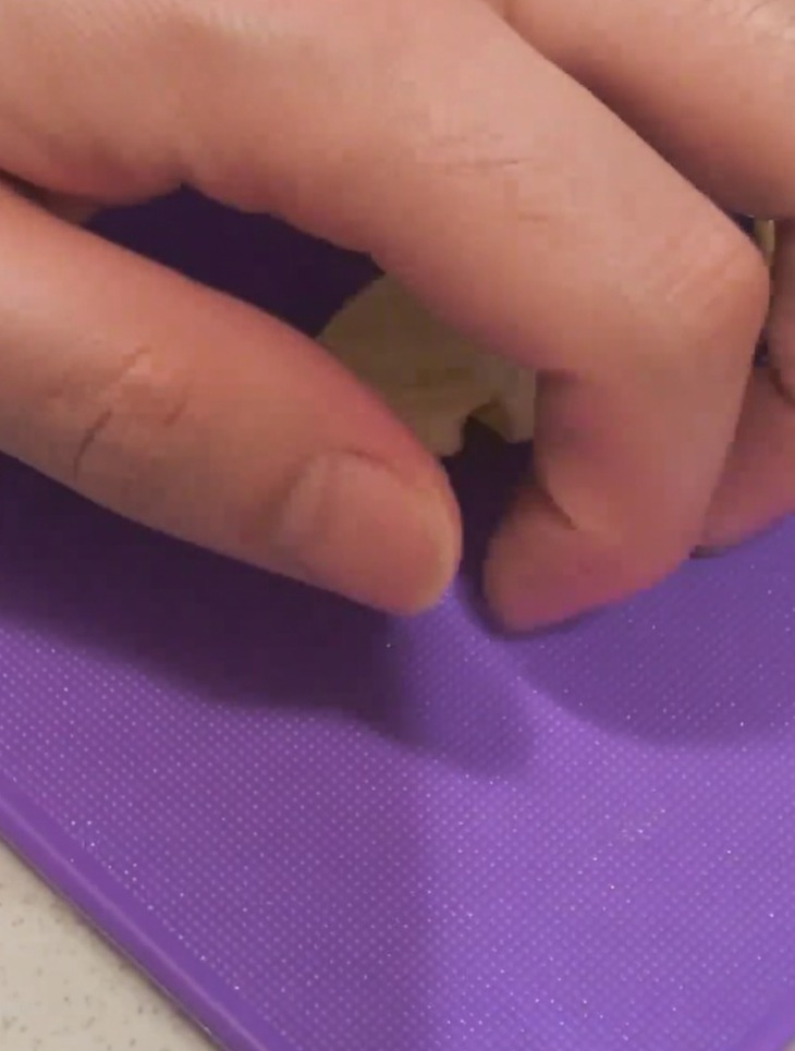 Close-up of a hand pressing the flat side of a knife onto a garlic clove on a purple cutting board.