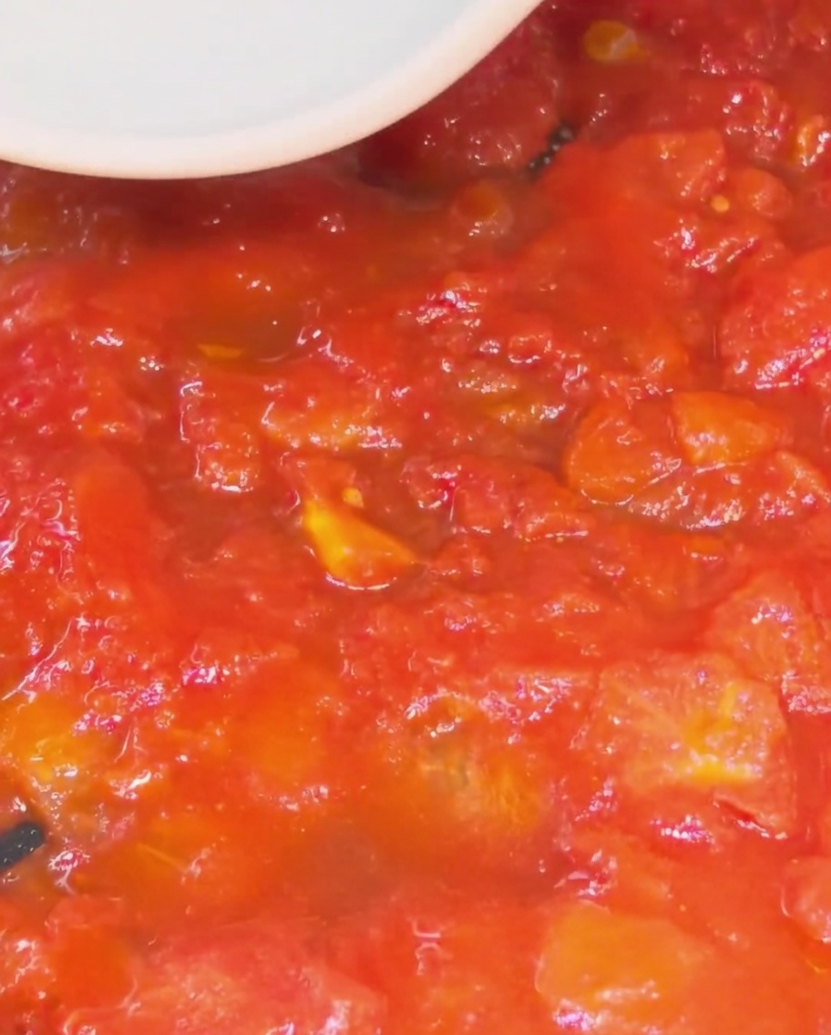 Close-up of a white silicone spatula pressing and mashing red tomato pieces into a thick, smooth sauce in a skillet.