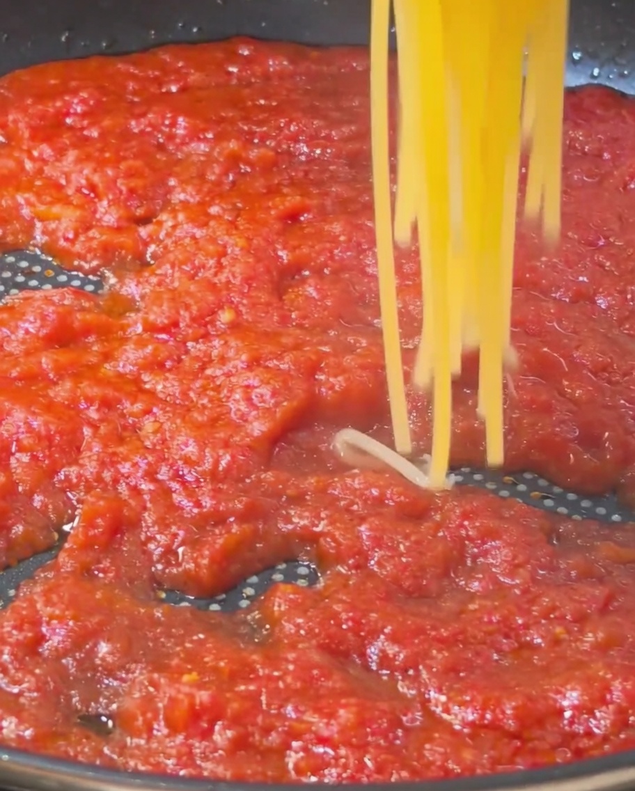 A portion of cooked golden spaghetti strands being lifted and placed into a pan filled with a rich, red tomato sauce.