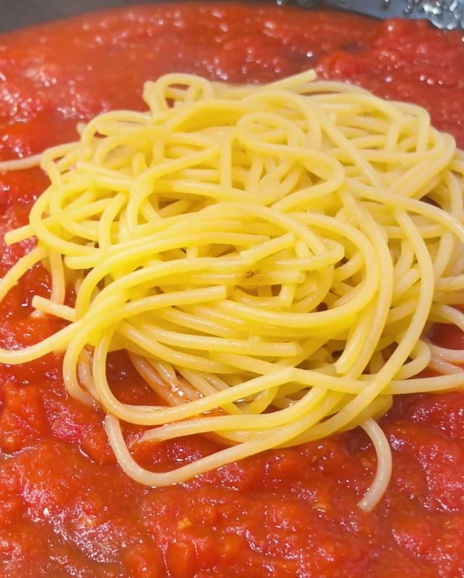 A spoonful of starchy pasta water being drizzled over a pile of spaghetti and tomato sauce in a pan.