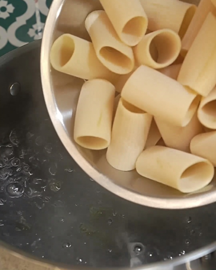 Dry rigatoni pasta being poured into a pot of vigorously boiling water.