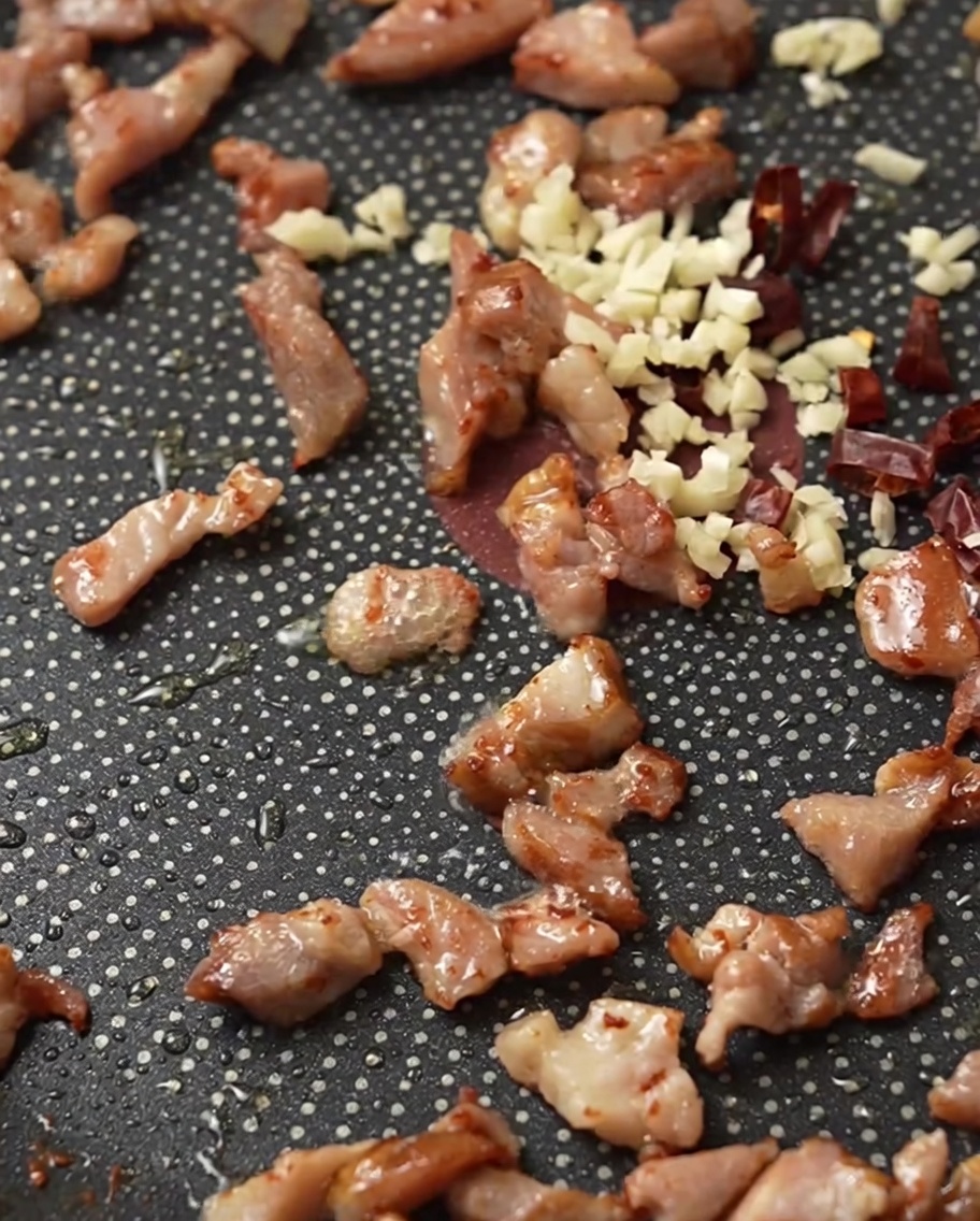 Diced bacon, minced garlic, and chopped dried red chilies sizzling in a frying pan.