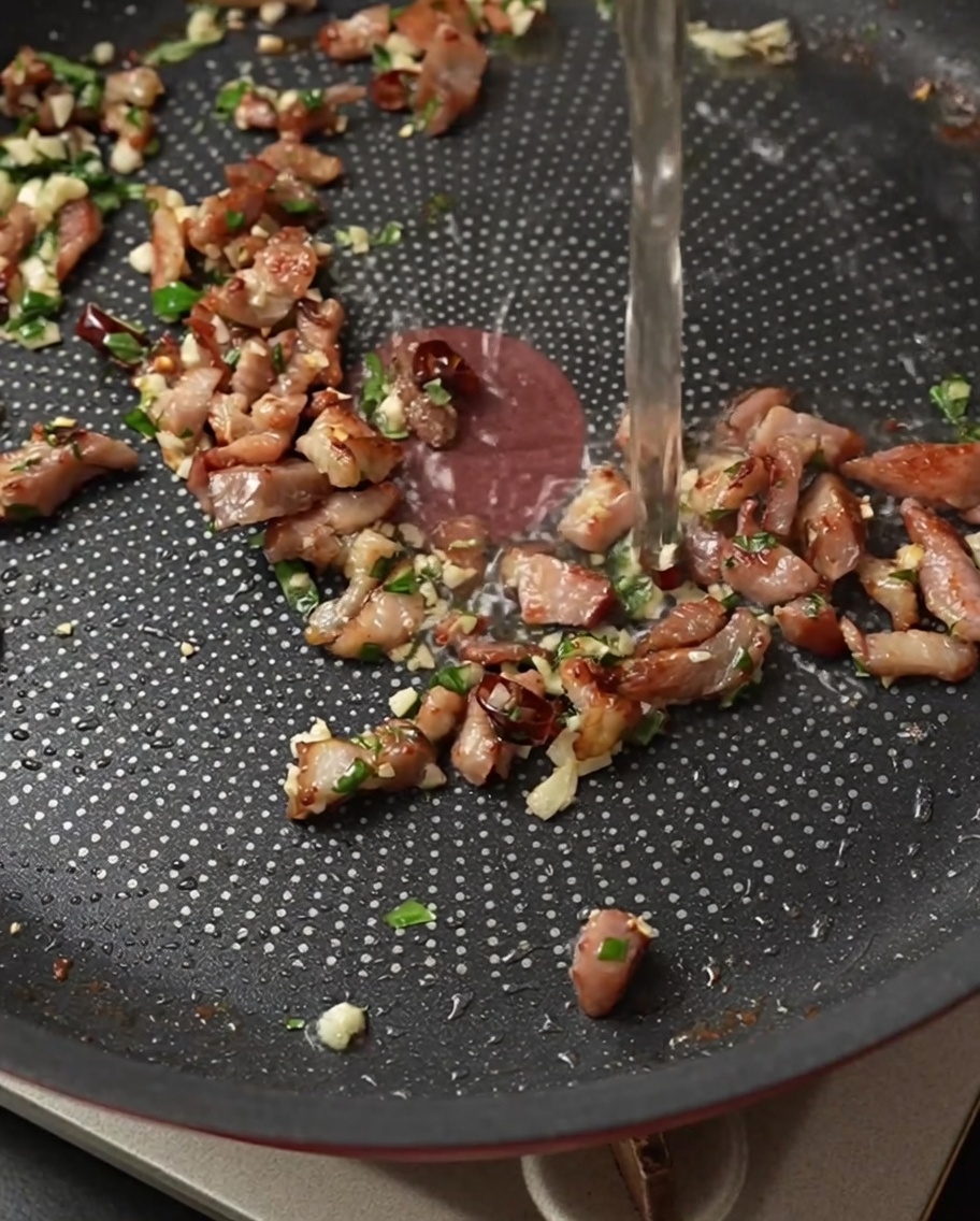 Clear white wine being poured into a pan filled with sizzling bacon, garlic, chili, and basil.
