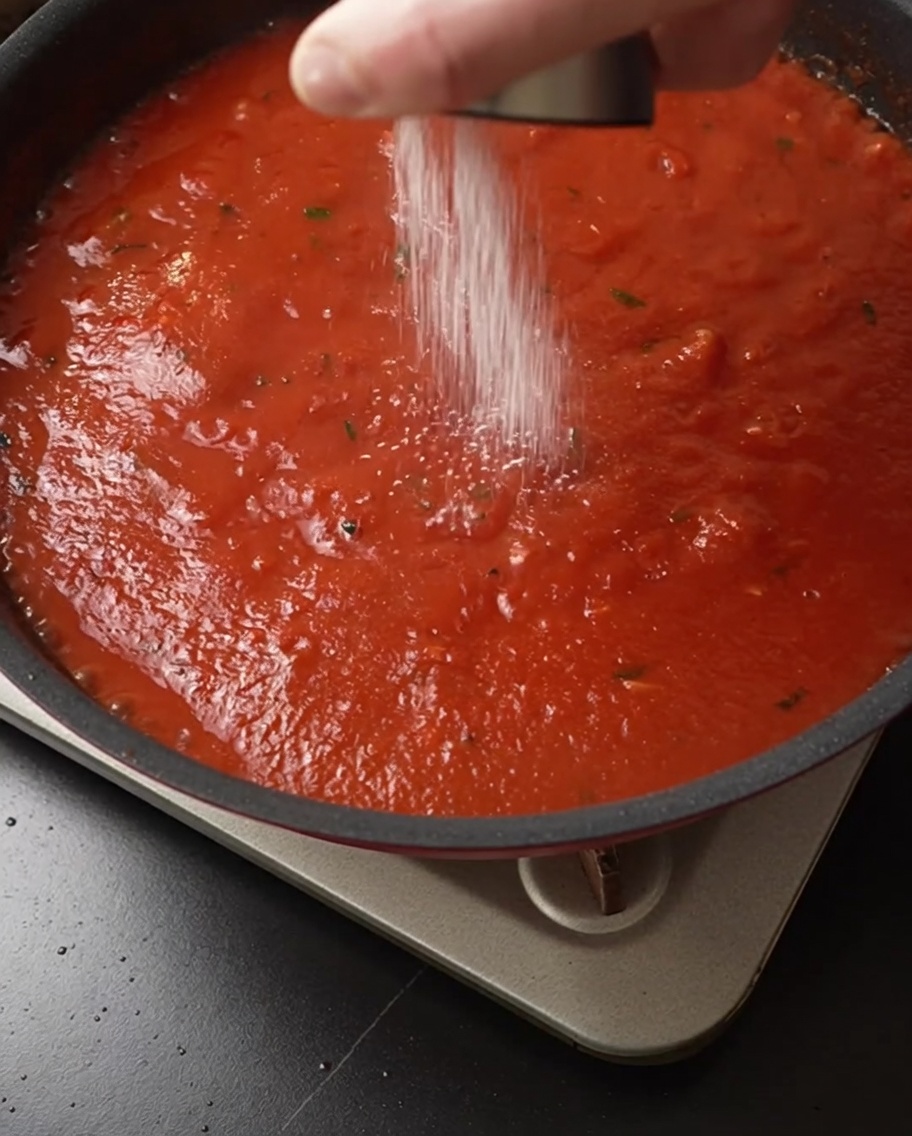 Salt being sprinkled from a shaker over a simmering, rich red tomato sauce in a pan.